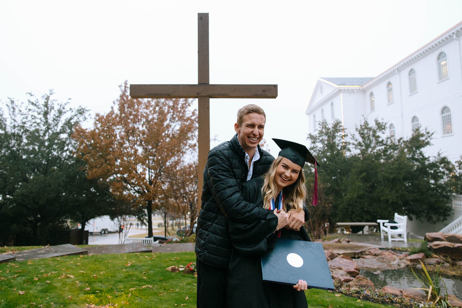 A young woman in graduation cap and gown holds her diploma while a young man hugs her from behind, both smiling, with a large wooden cross and a white building in the background