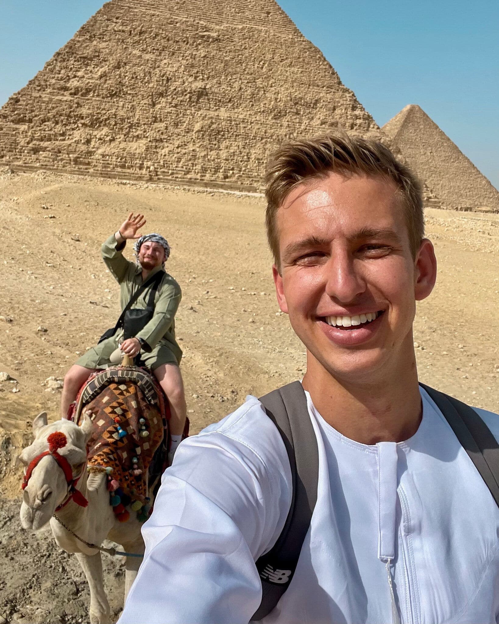 Two men posing for a selfie at the Pyramids of Giza, with one man riding a decorated camel in the background