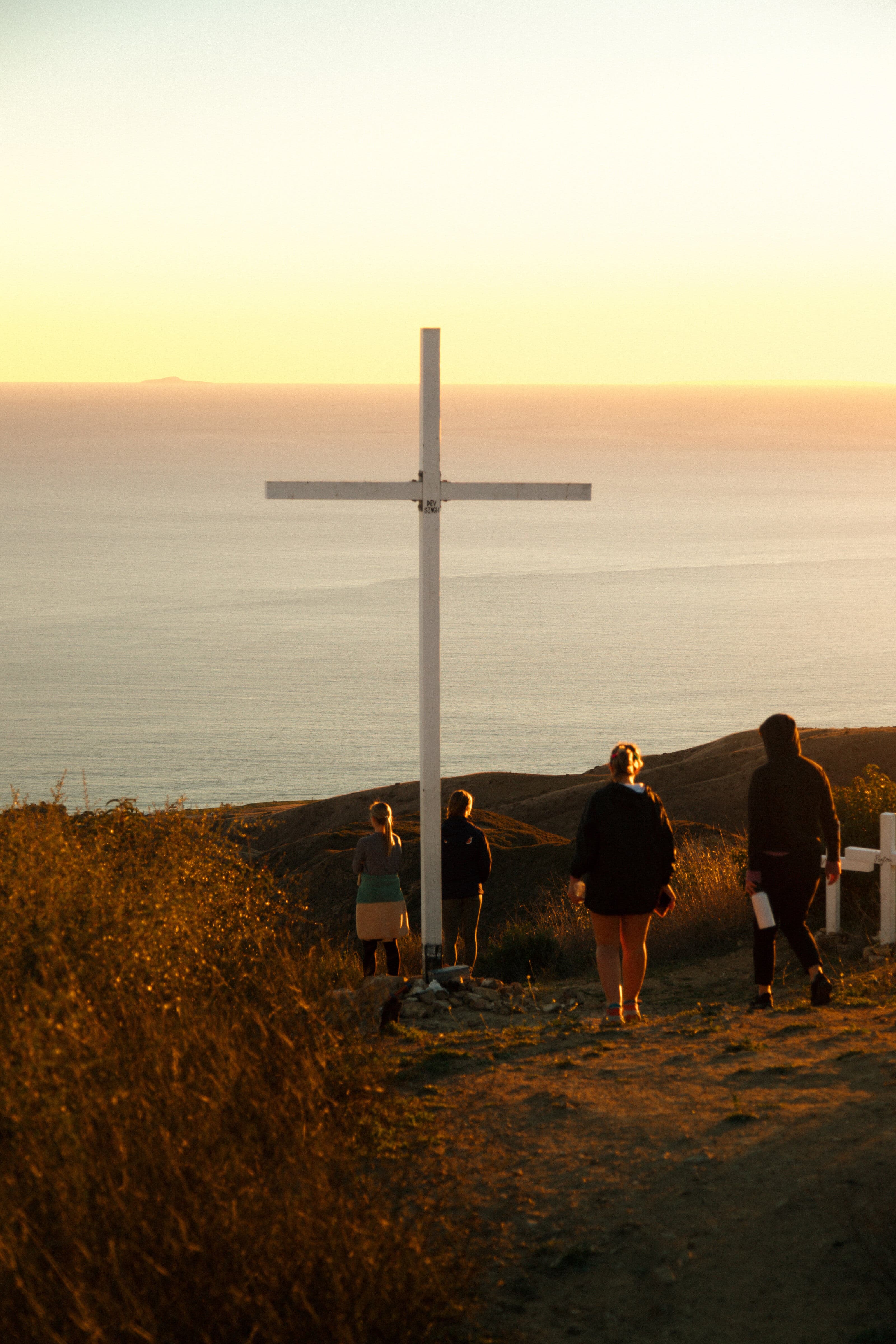 Four people gathered around a tall white cross on a coastal hillside at sunset, with the ocean visible in the background