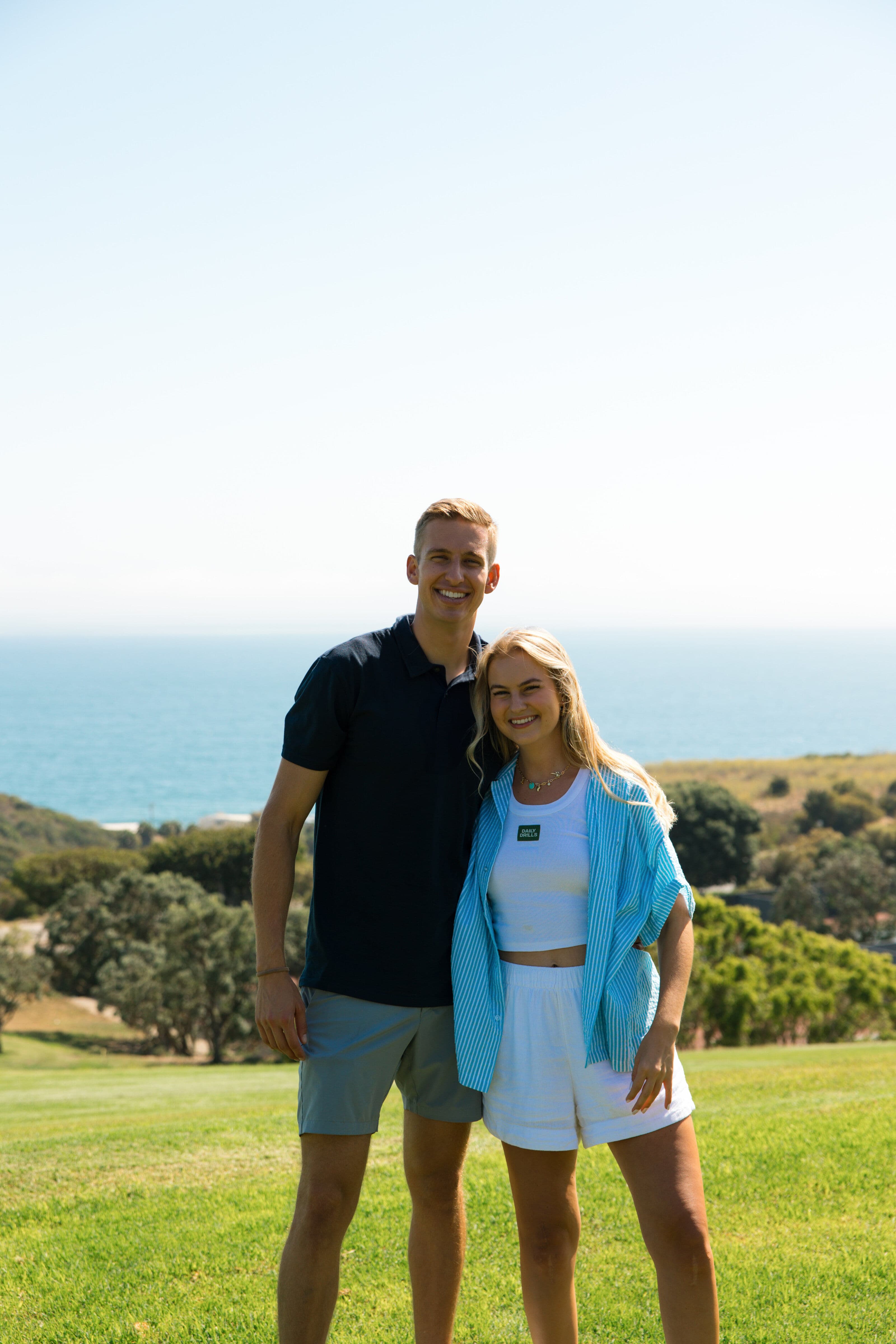 A smiling young couple standing together on a green golf course with an ocean view in the background
