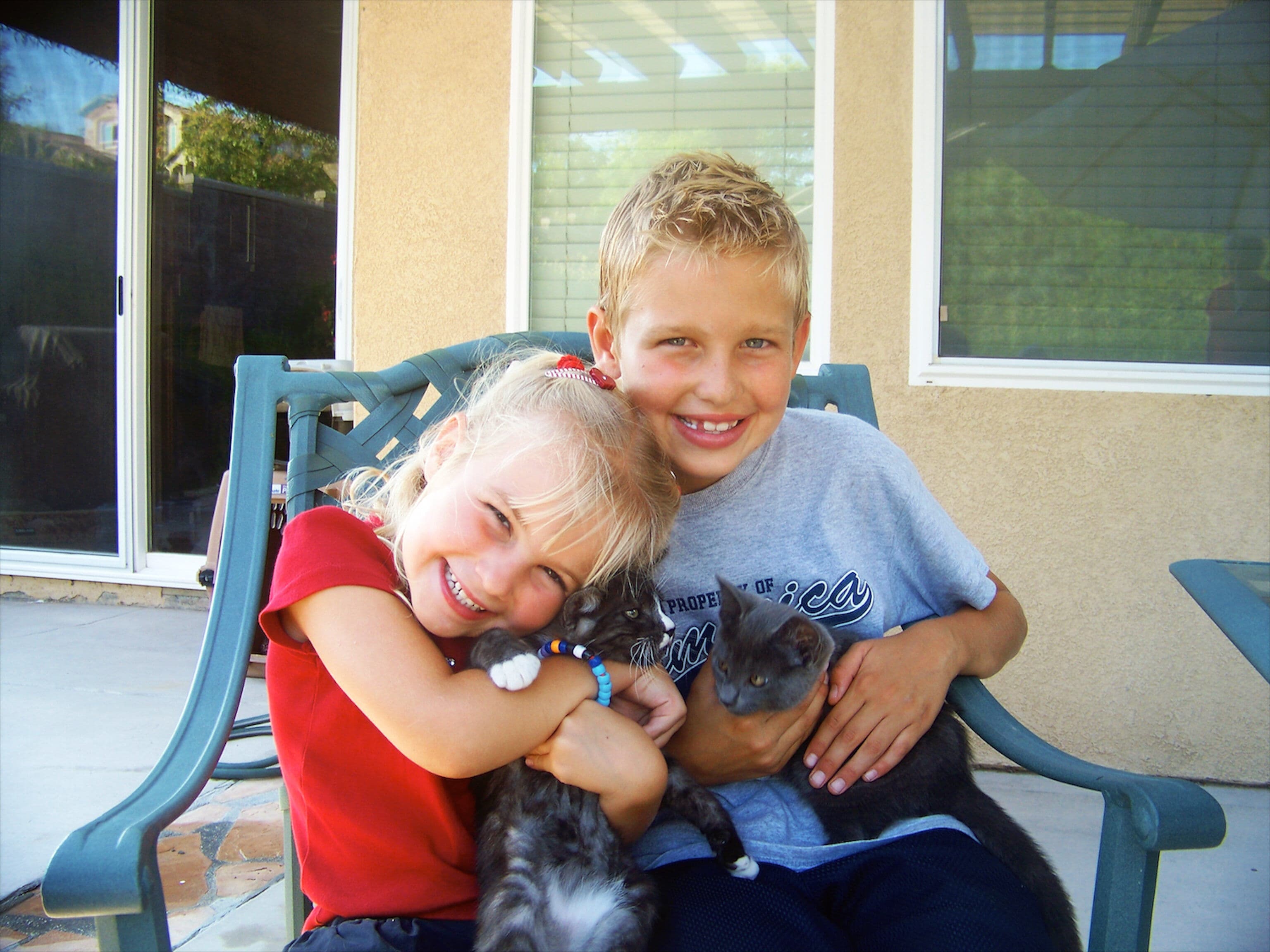 Two smiling blonde children sitting on a patio bench, each hugging a small gray kitten outside a suburban home