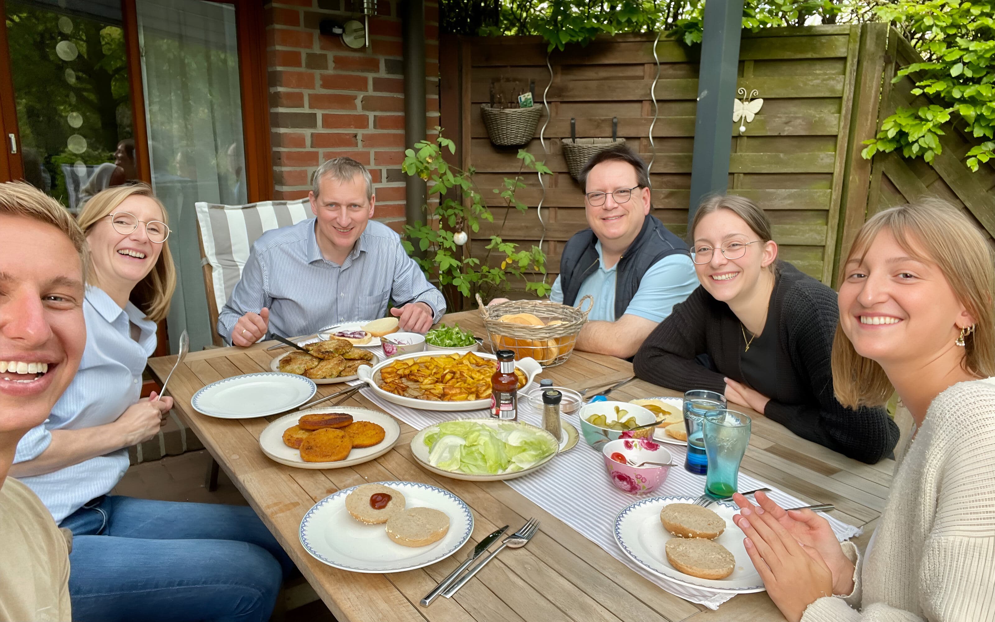 Six people smiling and sharing a burger and fries meal at an outdoor wooden patio table in a garden setting