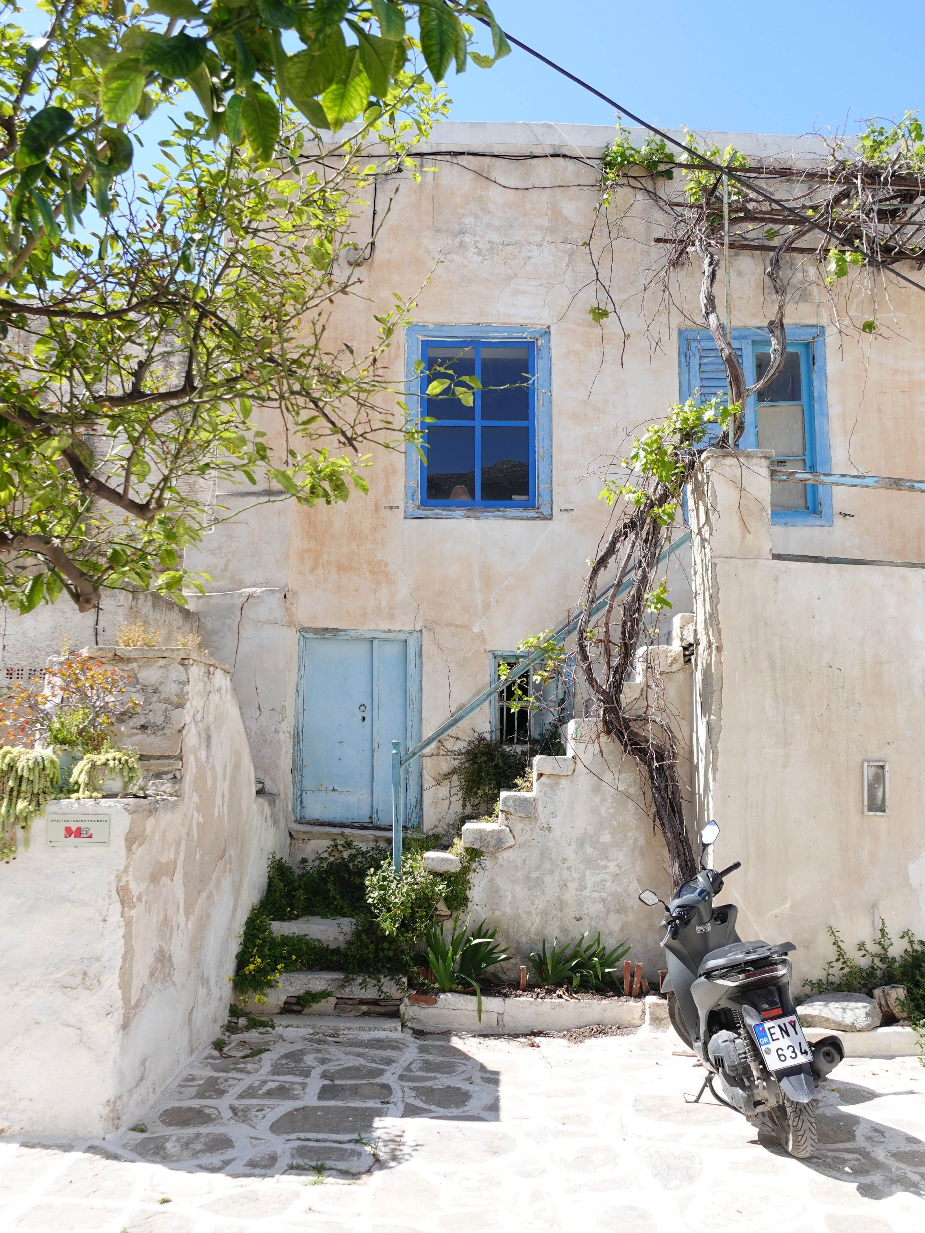 Weathered stone building with blue-painted windows and door, exterior staircase, and a scooter parked outside on a Greek island