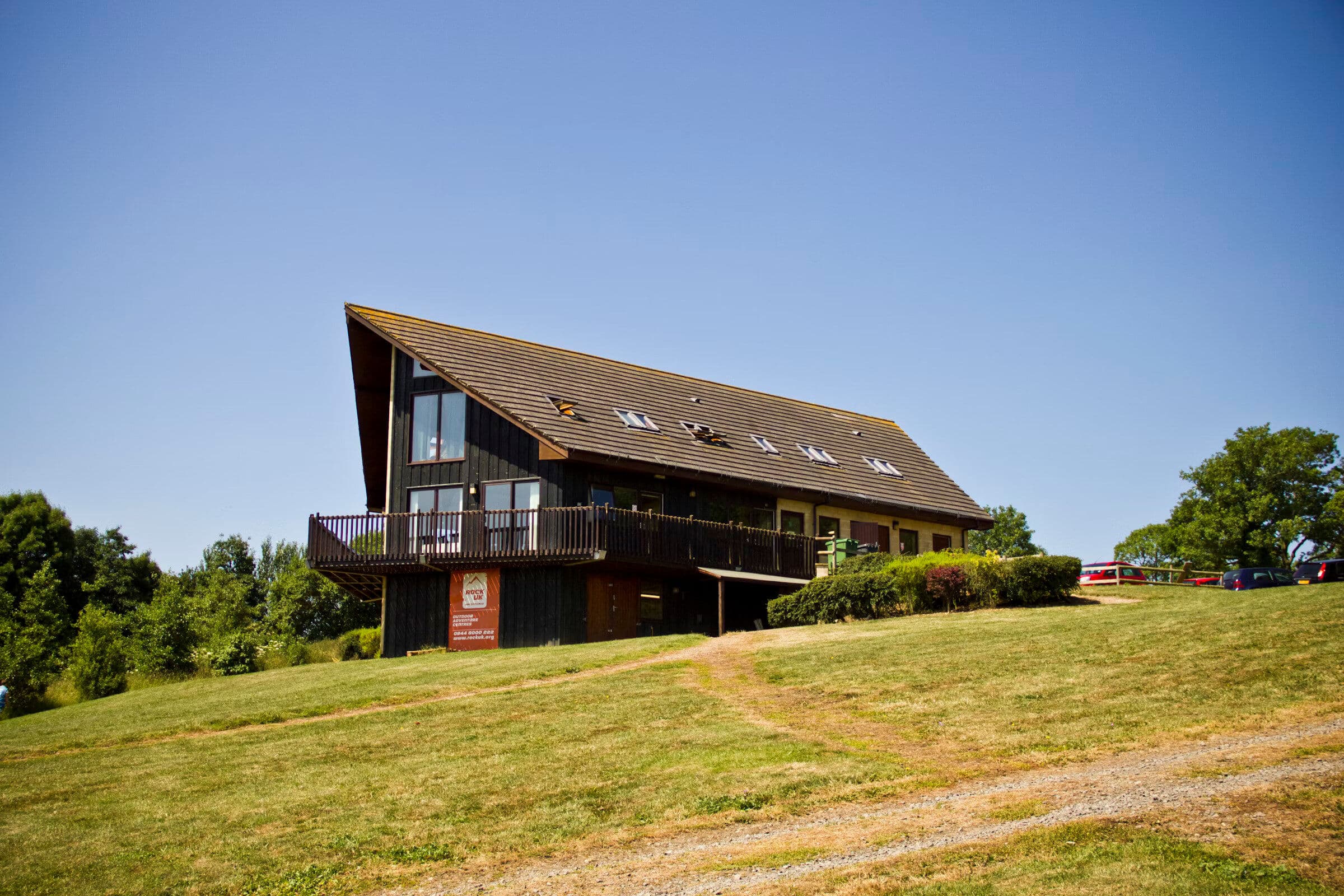 A large chalet-style building with a dramatic sloping roof and wooden deck, situated on a green hillside under a clear blue sky
