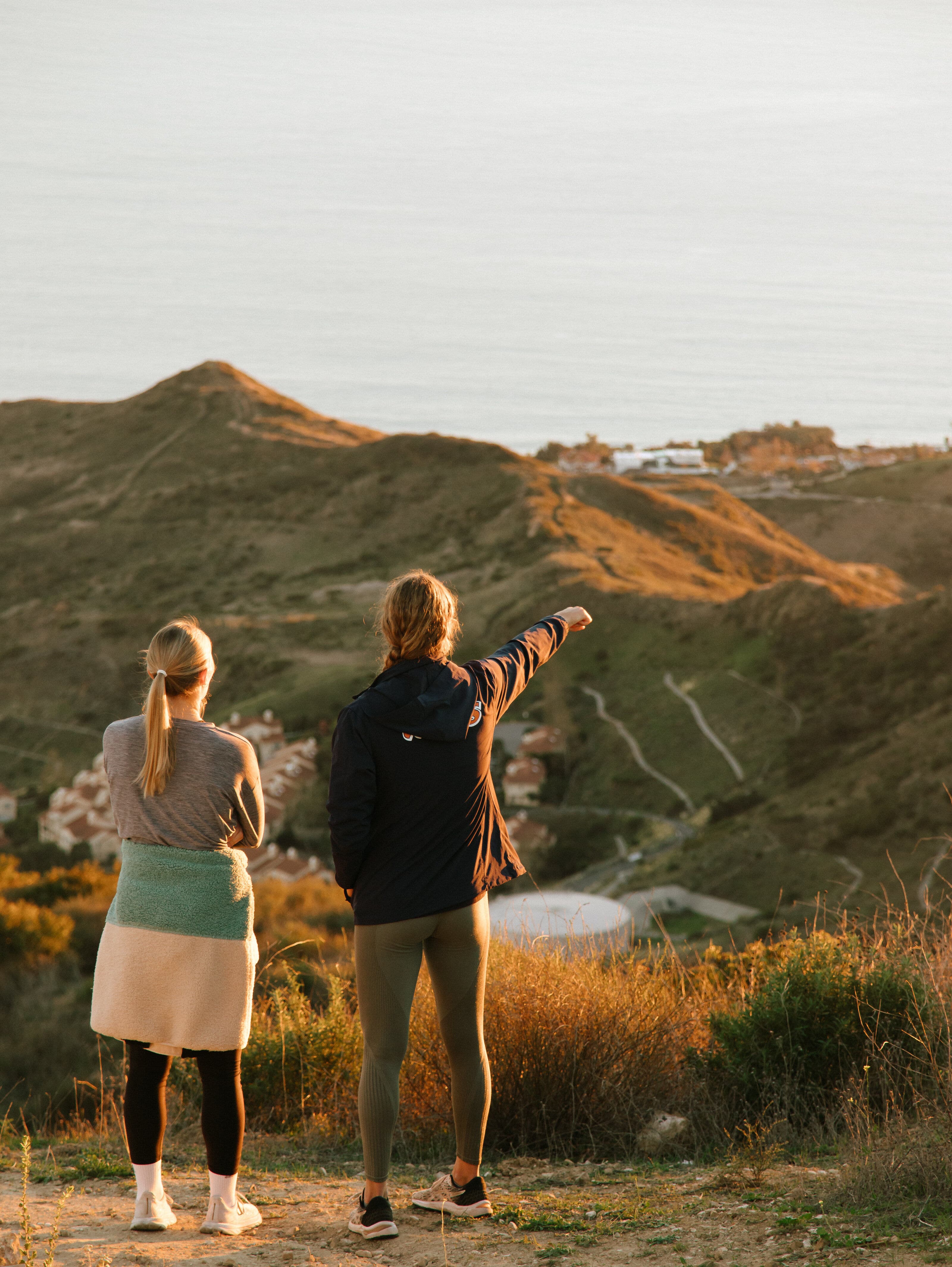 Two women standing on a hilltop at golden hour, one pointing toward rolling hills and a coastal town in the distance