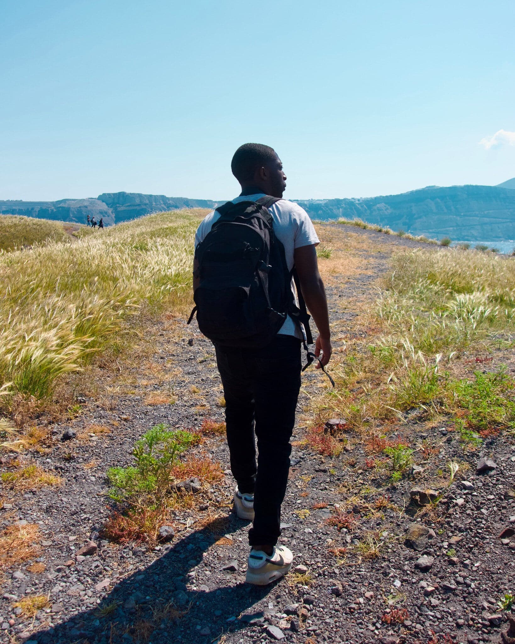 Young man with a black backpack walking along a rocky gravel trail through dry grasslands with mountains and sea in the distance