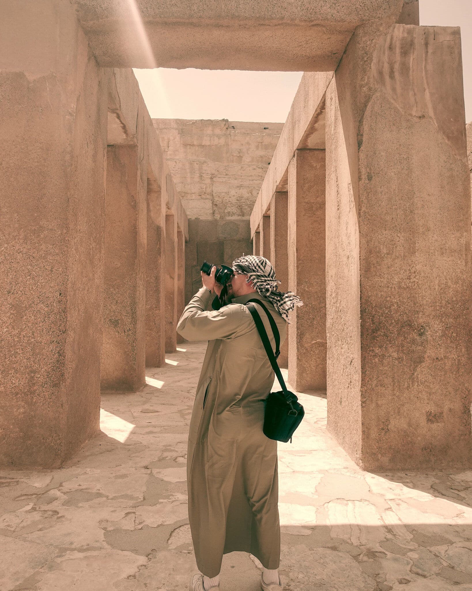 A person wearing a traditional thobe and keffiyeh photographs through a camera while standing in a narrow corridor between ancient stone pillars at an archaeological site