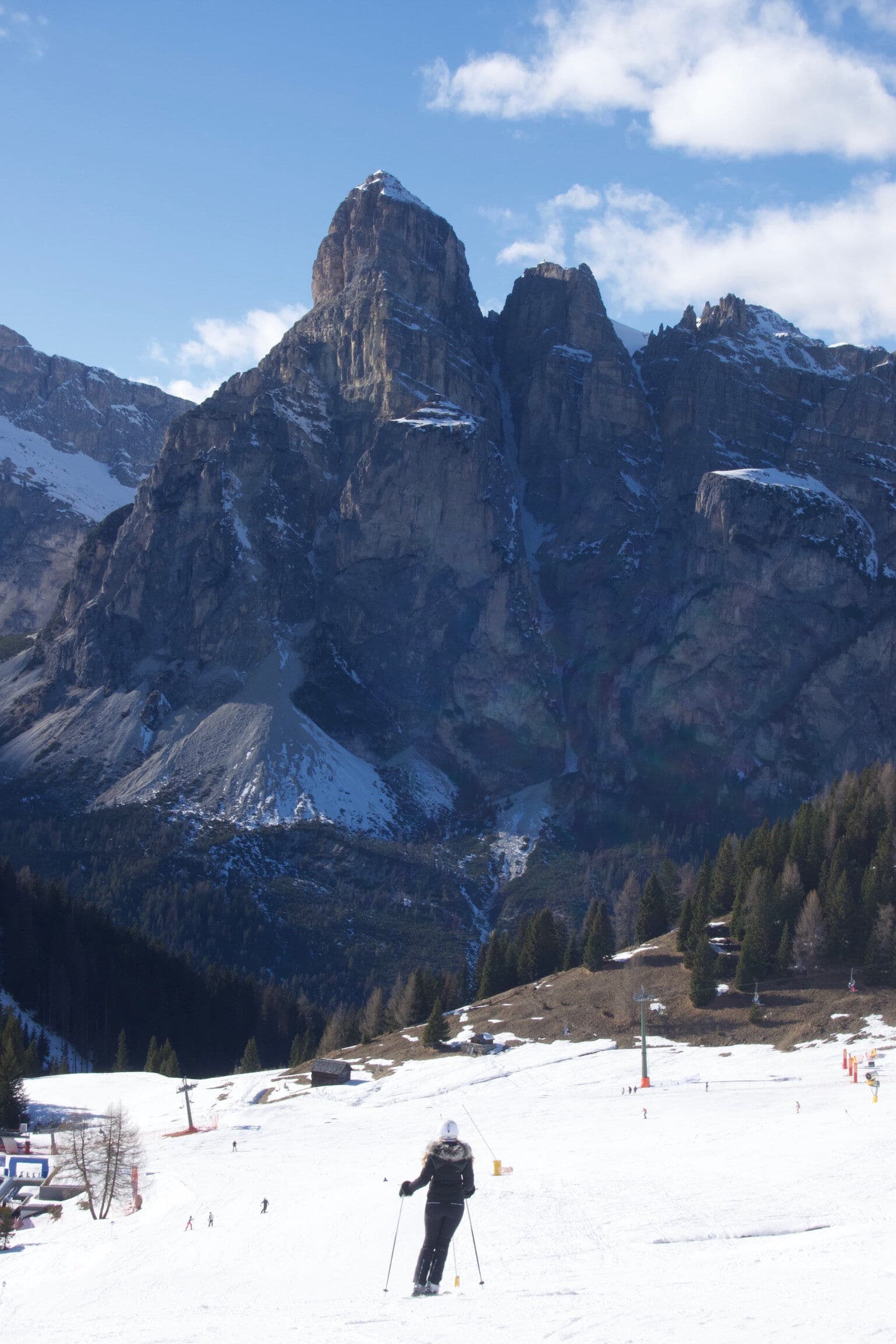 A skier stands on a snowy slope with dramatic rocky Dolomite peaks rising in the background under a blue sky